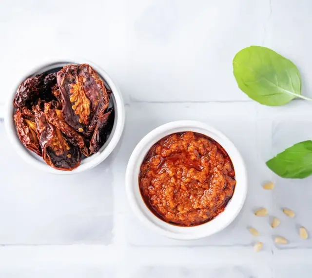 Due ciotoline bianche: una con pomodori secchi interi, l'altra con un invitante pesto di pomodori secchi. Foglie di basilico e pinoli completano la scena.