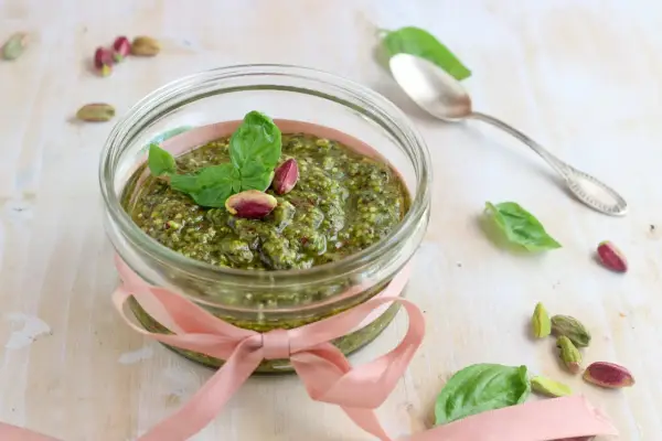 Un vasetto di pesto di pistacchi, decorato con un nastro rosa e guarnito con foglie di basilico e pistacchi interi, pronto per essere gustato.